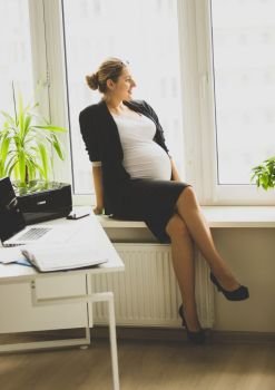 Young pregnant businesswoman sitting on windowsill and taking on phone