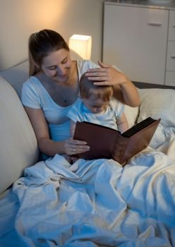 Beautiful smiling mother reading fairytale book to her baby at bed