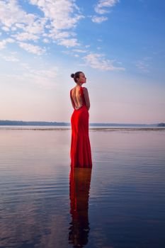 Attractive girl in a red dress standing in water on a background of beautiful sunrise. Beauty, fashion, outdoor
