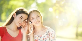 Two Smiling Young Girls with Pleasure Listening to Music on Headphones on a fine Summer Day