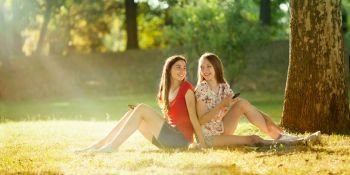 Two Beautiful Teenagers Holding Mobil Phones Sitting under a Tree in the Bright Summer Day