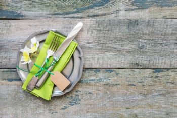 Ancient metal plate with a green napkin, vintage cutlery and spring flowers on the background of an old wooden table with space for text