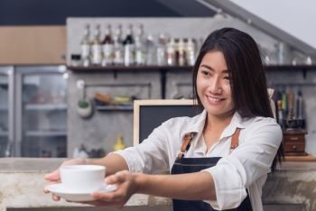 Close up of a young Asian female barista hold a cup of coffee serving to her customer with smile surrounded with bar counter background. Young female barista and her small shop. Food and drink concept