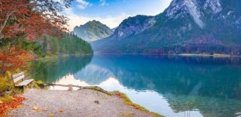 Contemplative scenery with a wooden bench placed on the Alpsee lake shore, in autumn decor, with the Bavarian forests and mountains reflected in the water.