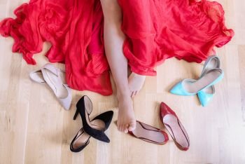 Above image with the bare feet of a Caucasian woman, dressed in a red dress, laying on the floor and surrounded by high heels shoes.