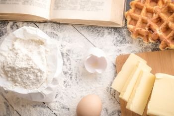 High angle view with a table full of wheat flour, sliced butter, egg shells, an open antique cookbook and freshly baked Belgian waffles.