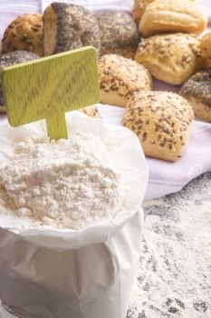 White paper bag full of flour with a wooden banner stick in it, homemade bread rolls decorated with seeds in the background on a kitchen towel.