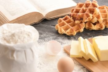 Delicious homemade Belgian waffles surrounded by the ingredients for their baking, flour, butter, eggs and an open recipe book, on a rustic table.