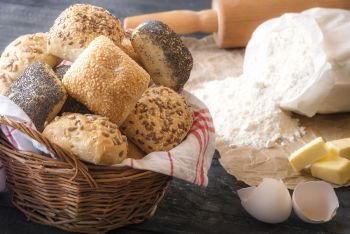 Baking theme image with a wicker basket full of homemade bread rolls, surrounded by flour, eggshell, butter and a rolling pin, on a rustic wooden table.