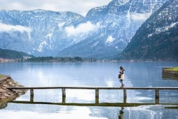 Travel destination theme image with a woman walking on a narrow deck that crosses the Hallstatter lake, surrounded by the Austrian Alps mountains.