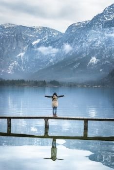 Famous places theme image with a woman standing on a deck, thinking and looking at the Austrian Alps and the Hallstatter lake, located in Hallstatt, Austria.