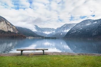 Holiday locations theme image with the Dachstein mountains reflected in the Hallstatter lake water and a wooden bench placed on its shore, in Hallstatt, Austria.