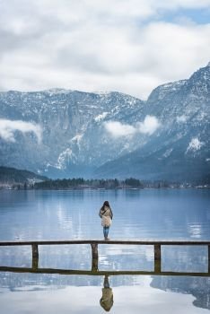 Famous places theme image with a woman standing on a deck, thinking and looking at the Austrian Alps and the Hallstatter lake, located in Hallstatt, Austria.