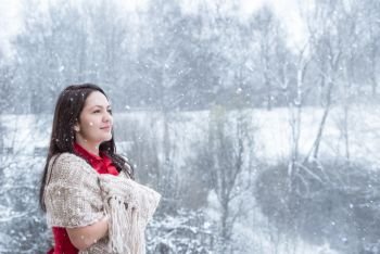 Attractive young woman with long dark hair, in a red dress, covered by a handmade shawl, smiling, looking up at the falling snowflakes.