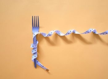  Diet and weight control concept. Measuring tape wrapped around fork lying on yellow background. 