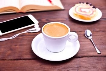cup of espresso coffee on a brown wooden background, behind a smartphone with headphones and an open book