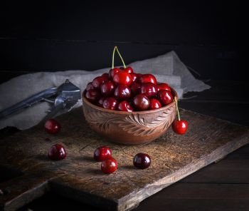ripe red cherry in a round clay bowl on a brown wooden board, black background. ripe red cherry in a round clay bowl 
