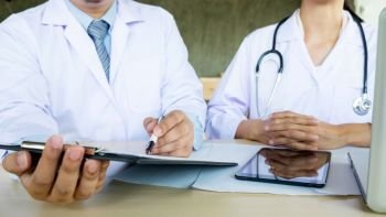 Two doctors discussing patient notes in an office pointing to a clipboard with paperwork as they make a diagnosis or decide on treatment.