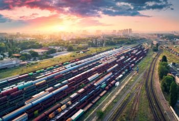 Cargo trains. Aerial view of colorful freight trains. Railway station. Wagons with goods on railroad. Heavy industry. Industrial scene with trains, city buildings and cloudy sky at sunset. Top view 