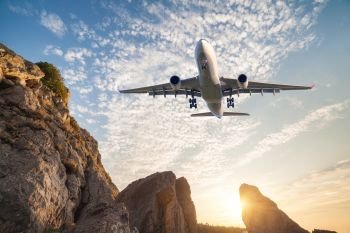 Big white aircraft is flying over rocks at sunset. Landscape with passenger airplane, mountains, colorful blue sky with clouds. Passenger airplane is landing. Business travel. Commercial plane.