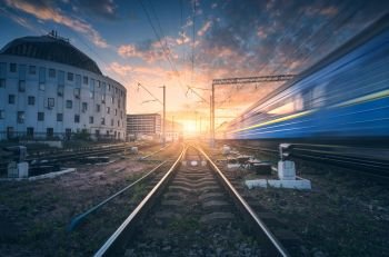 High speed passenger train in motion on railroad track at sunset. Railway station with blurred modern commuter train,  against colorful blue sky with red and orange clouds at dusk.Industrial landscape