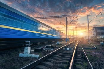 High speed passenger train in motion on railroad track at sunset. Railway station with blurred modern commuter train,  against colorful blue sky with red and orange clouds at dusk.Industrial landscape