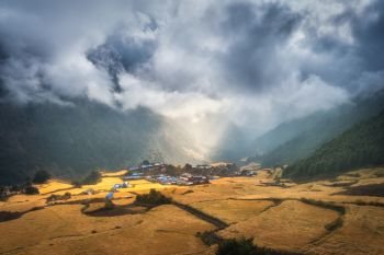 Amazing small village on the hill lighted by a sunbeam at sunrise. Landscape with houses, fields with yellow grass, yellow sunlight and mountains with green forest in Nepal. Rustic view. Himalayas