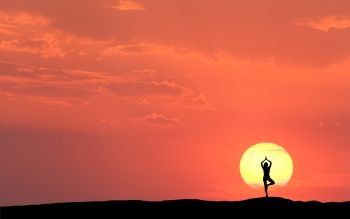 Silhouette of a standing sporty woman practicing yoga with raised up arms on the hill on the background of sun and colorful orange sky with clouds. Landscape with meditating girl at sunset. Fitness