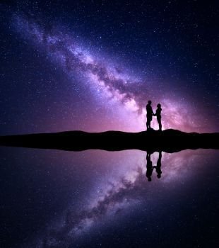 Milky Way with silhouette of people. Landscape with night sky. Milky Way with silhouette of people. Landscape with night starry sky and man and woman holding hands on the mountain near the lake with reflection in water. Hugging couple with milky way. Universe