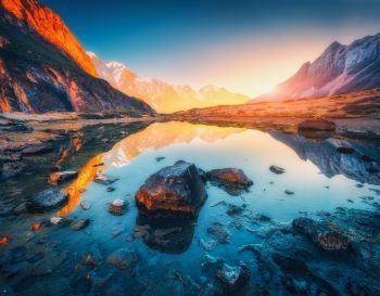 Beautiful landscape with high mountains with illuminated peaks, stones in mountain lake, reflection, blue sky and yellow sunlight in sunrise. Nepal. Amazing scene with Himalayan mountains. Himalayas. Mountains with illuminated peaks, stones in mountain lake at sun
