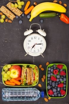 Sandwich, apple, grape, carrot, berry in plastic lunch boxes, alarm clock and bottle of water on black chalkboard. Back to school concept.