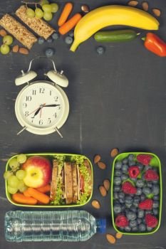 Sandwich, apple, grape, carrot, berry in plastic lunch boxes, alarm clock and bottle of water on black chalkboard. Back to school concept.