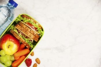 Green school lunch box with sandwich, apple, grape, carrot and bottle of water close up on white wooden  background. Healthy eating habits concept. Flat lay composition (from above, top view).