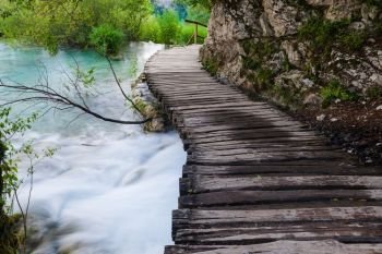 Wooden path across beautiful lake and waterfall in sunny green forest