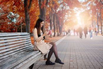 Beautiful girl holding cellphone and coffee sitting on the bench at city autumn park