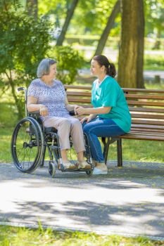 Caregiver talking to disabled senior woman in wheelchair outdoors in summer park. 