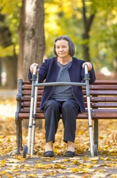Senior woman with walker getting up from bench and walking outdoors in autumn park. 