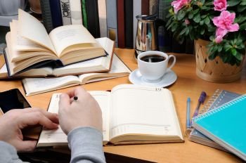 a man holds a pen and takes notes in a notebook on a desk in his office, surrounded by books, notebooks with a cup of black coffee