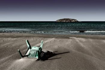 View of a small island from the Israeli shore of the Mediterranean Sea. Plastic chair buried in the sand of beach in Israel.