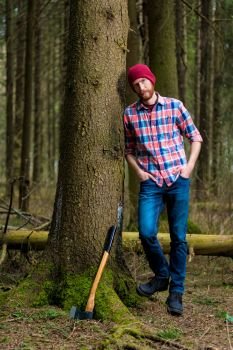 relaxed forester resting with an ax, leaning against a tree