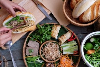 Woman hand make banh mi thit or Vietnamese bread, famous street food from  raw material: pork, ham, pate, egg and fresh herbs as scallions, coriander, carrot, cucumber, chilli.