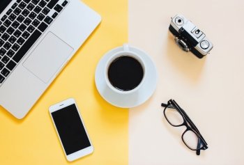 Creative flat lay photo of workspace desk with laptop, smartphone, coffee, eyeglasses and film camera on yellow modern background
