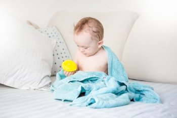 Adorable baby boy covered in blue towel playing with yellow rubber duck on sofa after bathing