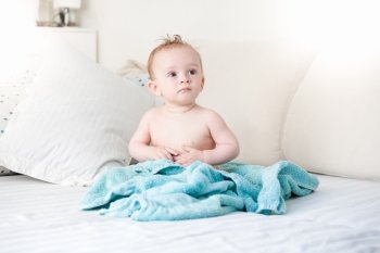 Freshly bathed baby boy covered in blue towel sitting on big sofa at living room