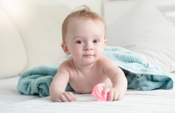 Portrait of adorable baby lying under blue towel on bed and holding plastic toy