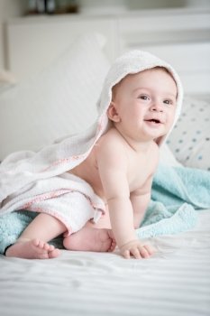 Beautiful smiling 9 months old baby boy sitting on bed covered in towel after bathing