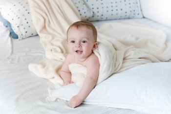 Portrait of cheerful baby boy lying on big pillow under white blanket
