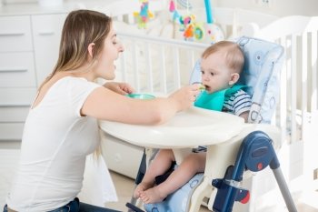 Beautiful smiling woman feeding her baby boy in highchair at living room