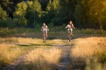 Toned image of two teenage girls riding bicycles on meadow at sunset