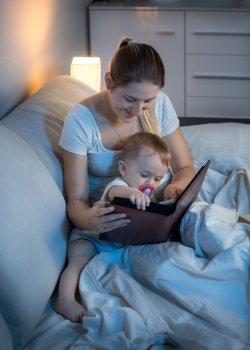 Portrait of beautiful young mother reading book to her baby in bed at night